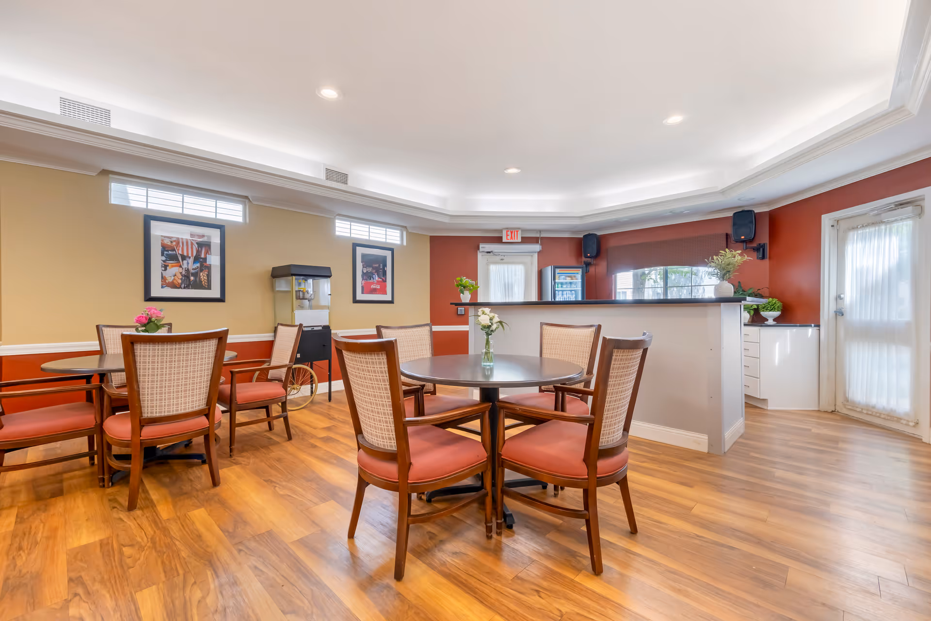 Bright communal dining room with round tables and chairs, a small service counter/bar, a popcorn machine and wood floors.