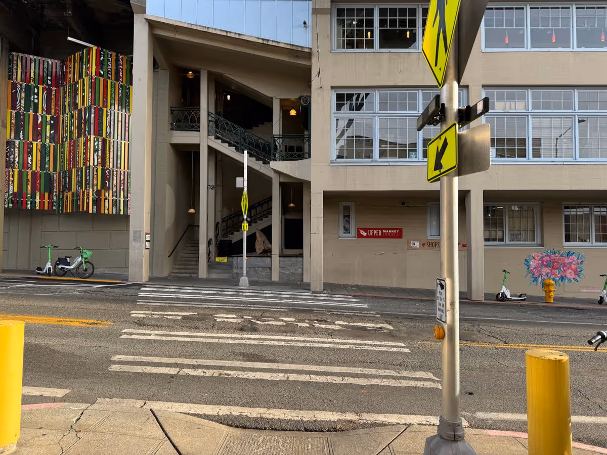 Exterior view of a multi-story building with an open stairwell, crosswalk, parked electric scooters, and colorful murals.