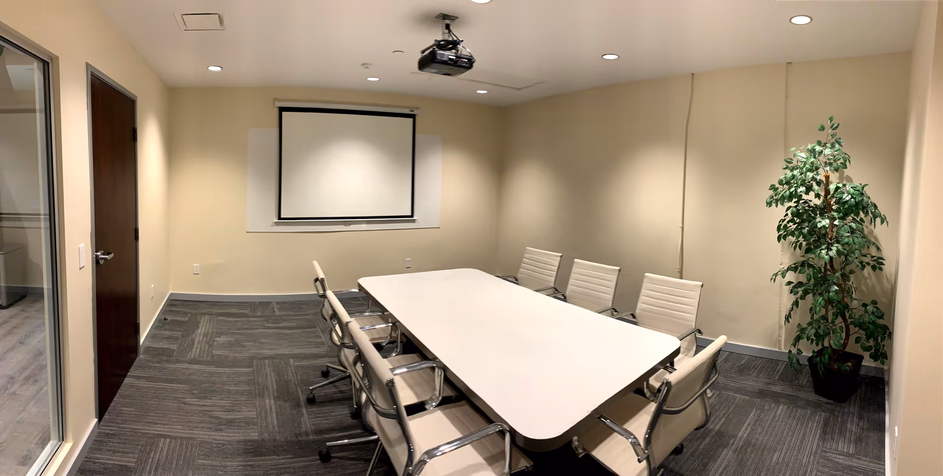 A conference room with a long table, white chairs, projector, screen, and potted plant.