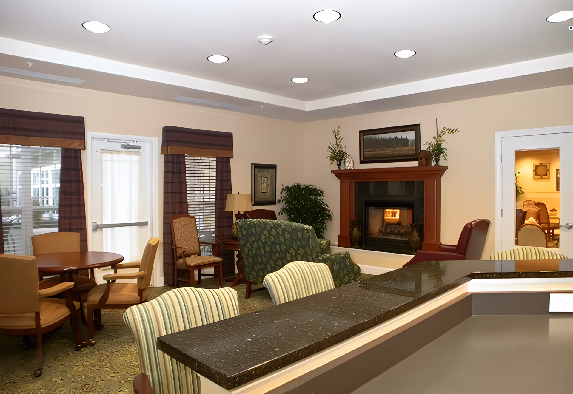 A cozy common area in a senior living facility featuring a fireplace with a wooden mantel, a green patterned armchair, a maroon armchair, and a round wooden table with beige chairs. The room has beige walls, recessed ceiling lights, and windows with brown valances. There is also a countertop with striped chairs in the foreground and a door leading to another room with more chairs visible.
