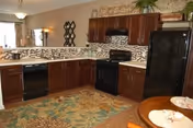 Interior view of a kitchen featuring dark wood cabinets, black appliances including a refrigerator, stove, and dishwasher, a tiled backsplash, and a patterned rug on the floor. A small round dining table with place settings is partially visible in the foreground.