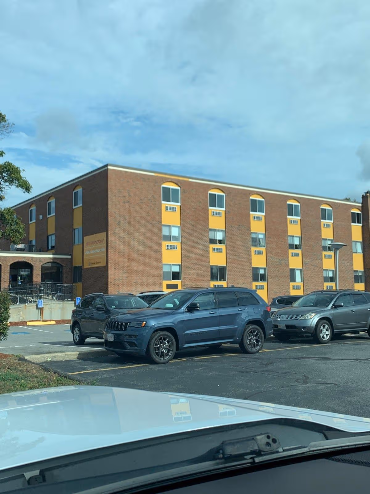 Brick senior living facility with yellow accents and parked cars seen from a car in lot.