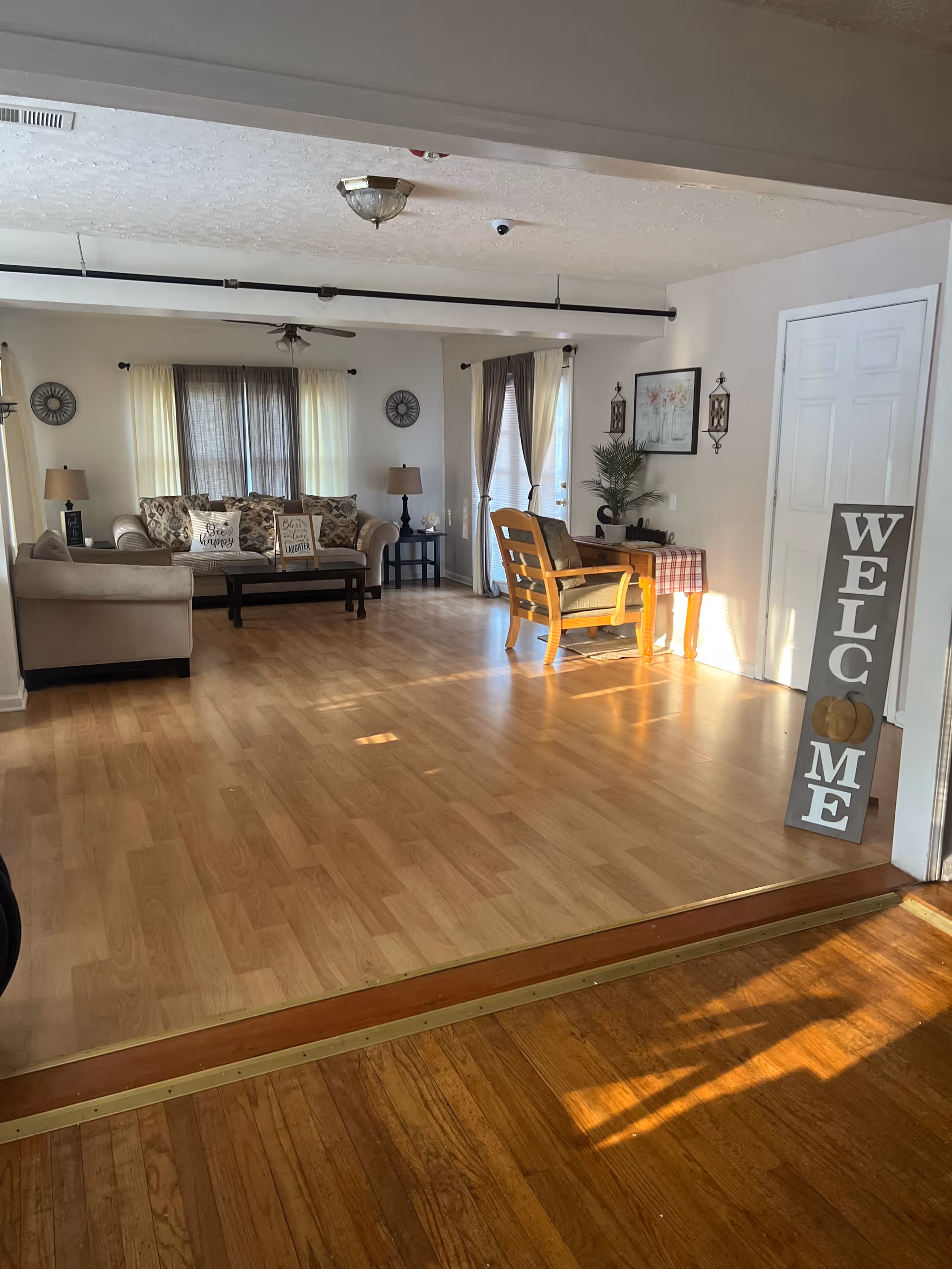 Spacious common living room with couches, chairs, lamps, and a welcoming sign by the entrance.