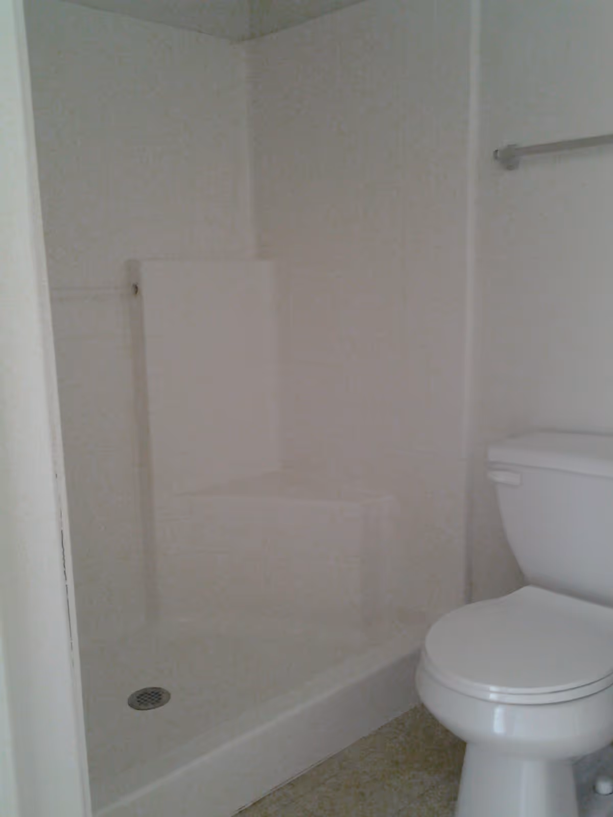 A small bathroom featuring a white toilet and a white walk-in shower with a built-in seat and a metal drain on the floor. There is a towel bar mounted on the wall above the toilet.