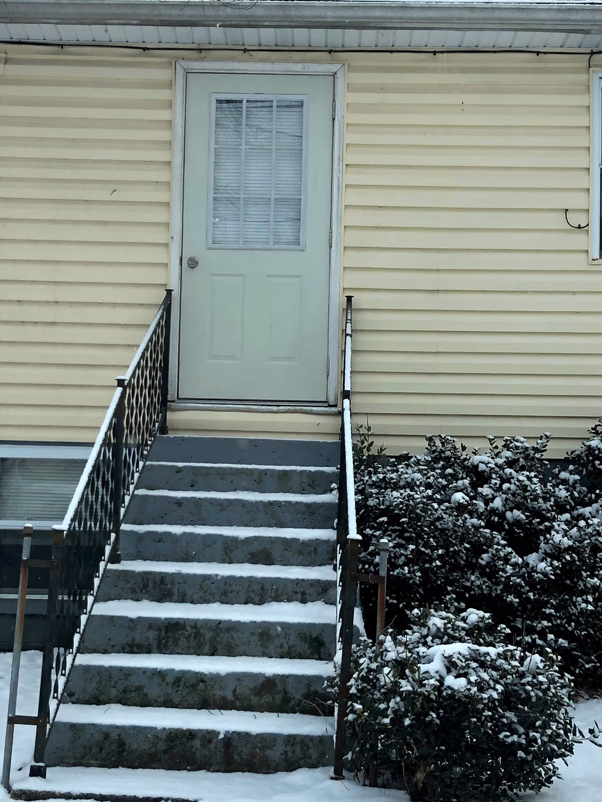 Yellow siding assisted living building entrance with snow-covered concrete steps and railing.