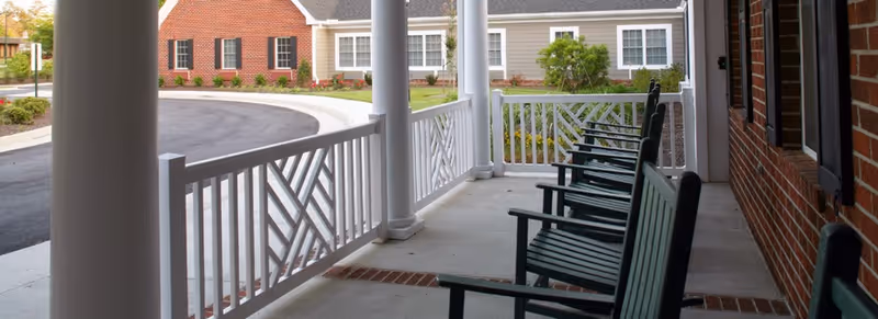 Covered front porch with a row of green rocking chairs overlooking a circular driveway and brick-and-siding buildings.