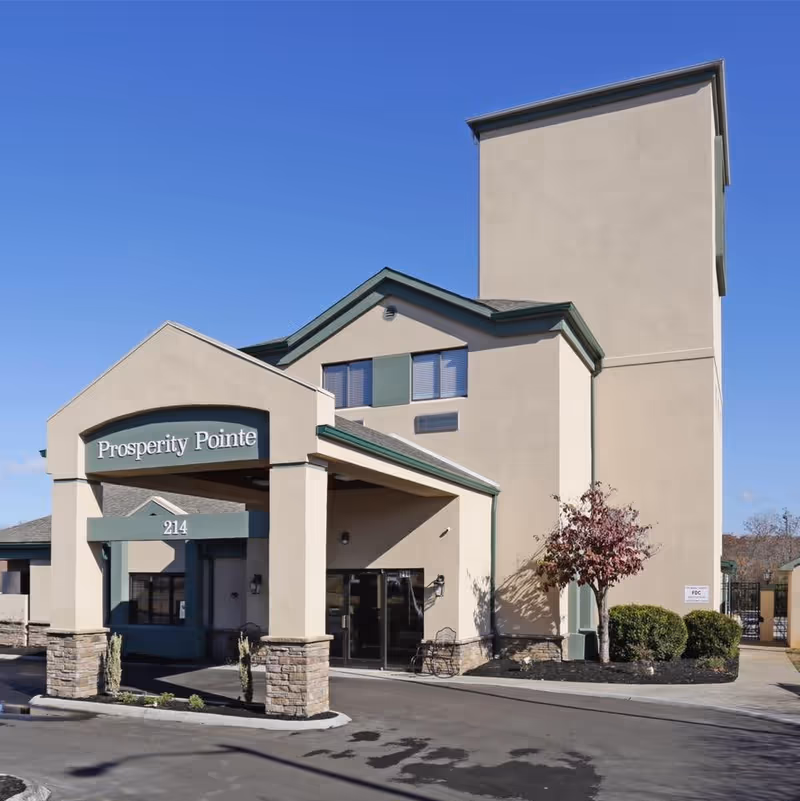 Exterior view of the Prosperity Pointe facility showing the entrance with a covered drop-off area, beige walls with green trim, stone pillars, and a clear blue sky.