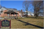 Brick retirement community building with a wooden gazebo, sign, and leafless trees on the lawn.