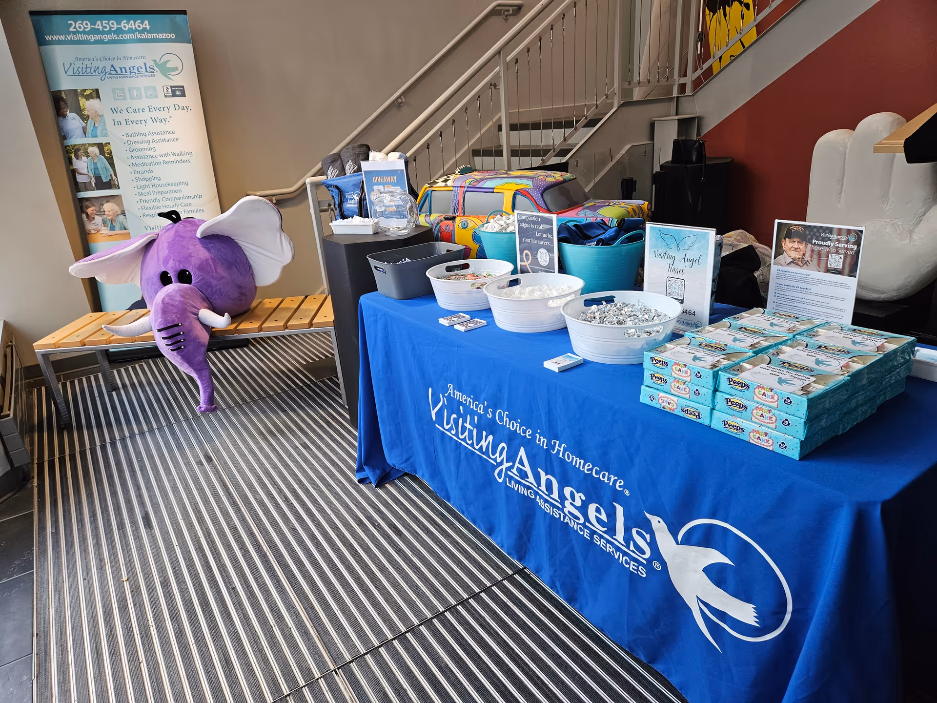Indoor promotional table with blue Visiting Angels tablecloth, brochures, and a purple elephant mascot near stairs.