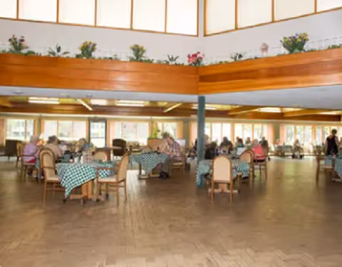 Interior dining hall with round tables, checkered tablecloths, and residents seated.