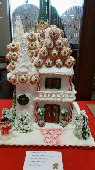 A festive gingerbread house decorated with candy and snowy icing on a red tablecloth inside a building.