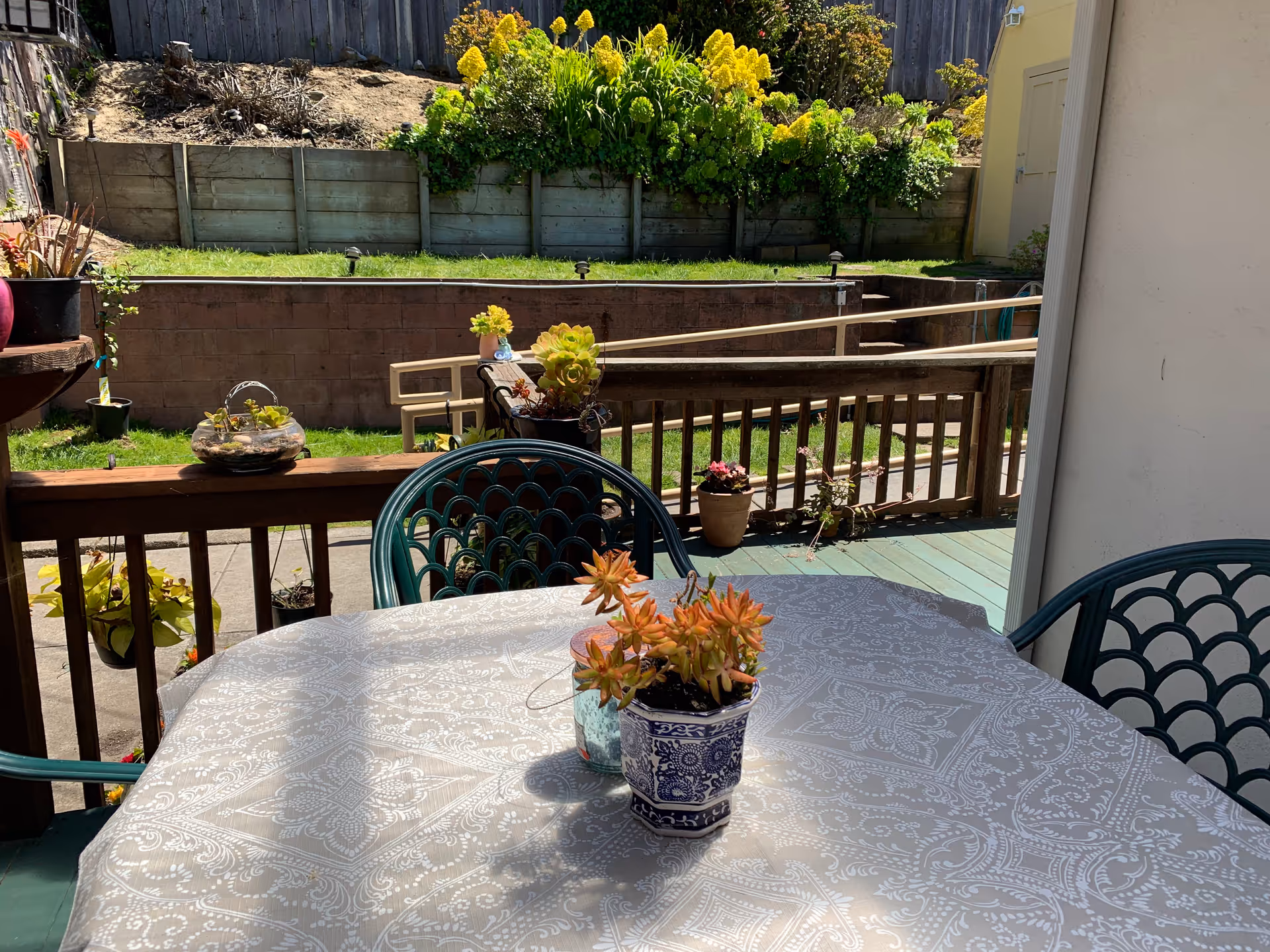 Outdoor patio with round table, patterned tablecloth, potted plants, and garden railing.