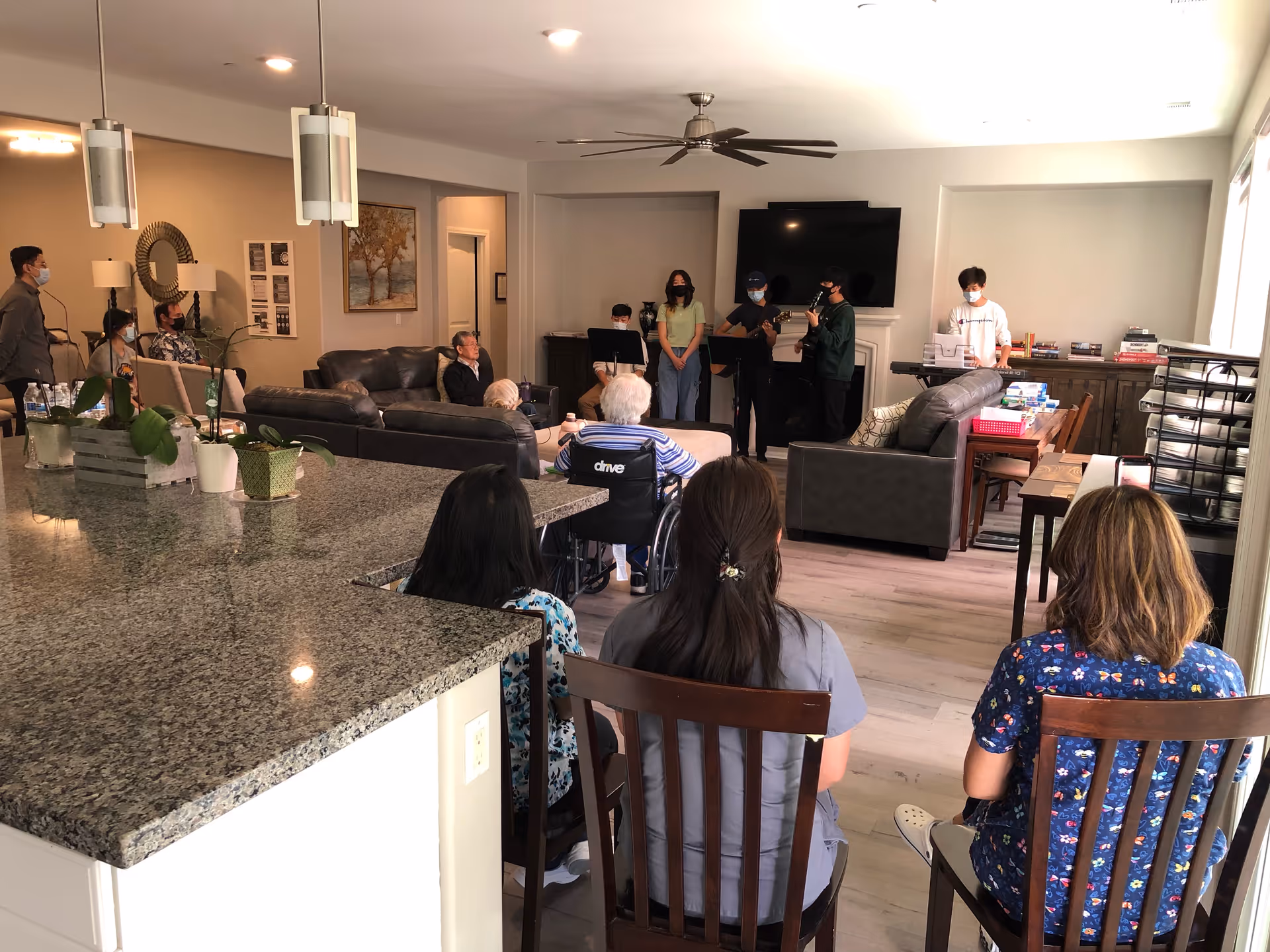 A group of elderly residents and staff seated in a living room area watching a small group of young musicians performing near a television mounted on the wall. The room has comfortable seating, a ceiling fan, and a kitchen counter with plants in the foreground.