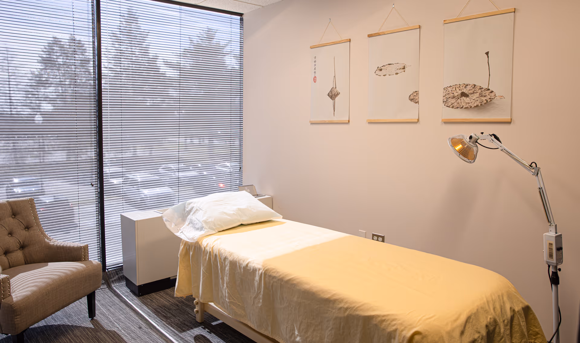 A bedroom-like clinic room with a single bed, chair, blinds, and wall art.