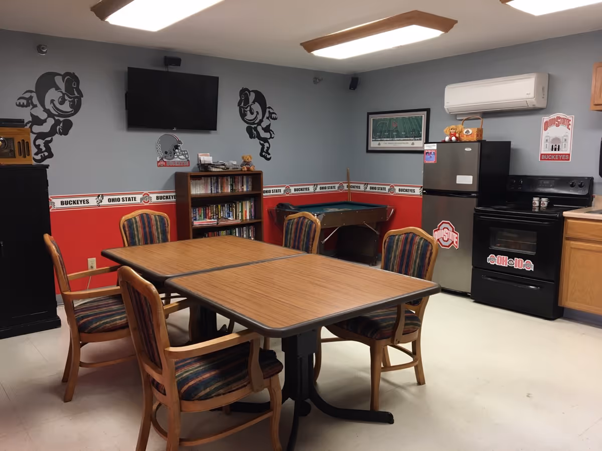 A room with a wooden table and six striped cushioned chairs around it. The walls are decorated with Ohio State Buckeyes themed decals and borders. There is a bookshelf filled with books, a small pool table, a wall-mounted TV, a refrigerator, and a stove. The room has a cozy, recreational atmosphere.