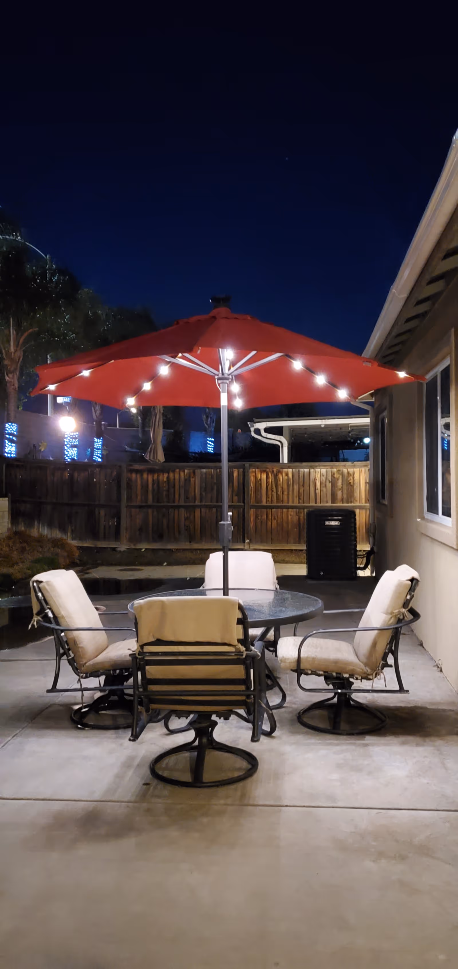 Outdoor patio area at night with a round glass table surrounded by four cushioned chairs. A large red umbrella with string lights is positioned in the center of the table. The area is enclosed by a wooden fence and adjacent to a building wall with a window.