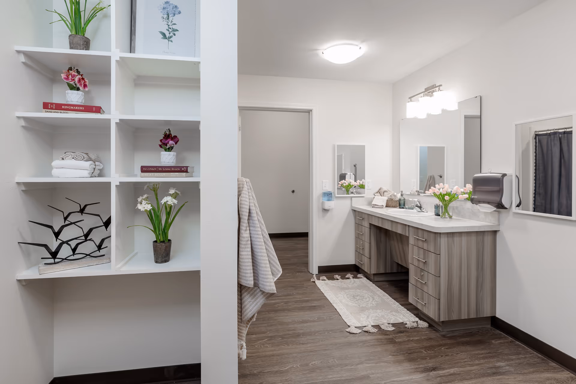 A clean and modern bathroom with a wooden vanity featuring multiple drawers, a large mirror, and a countertop with a sink. There is a vase with pink tulips on the countertop, a towel rack with beige towels, and a white decorative rug on the wooden floor. To the left, there is a white built-in shelving unit with decorative items including potted plants, books, and a black metal sculpture. The walls are white and the lighting is bright.