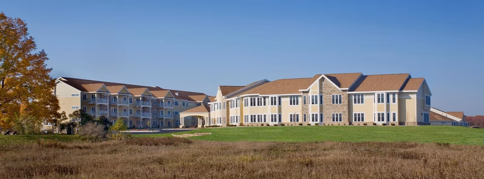 Exterior view of a large senior living facility building with beige siding and brown roofs, surrounded by green grass and trees with autumn foliage under a clear blue sky.