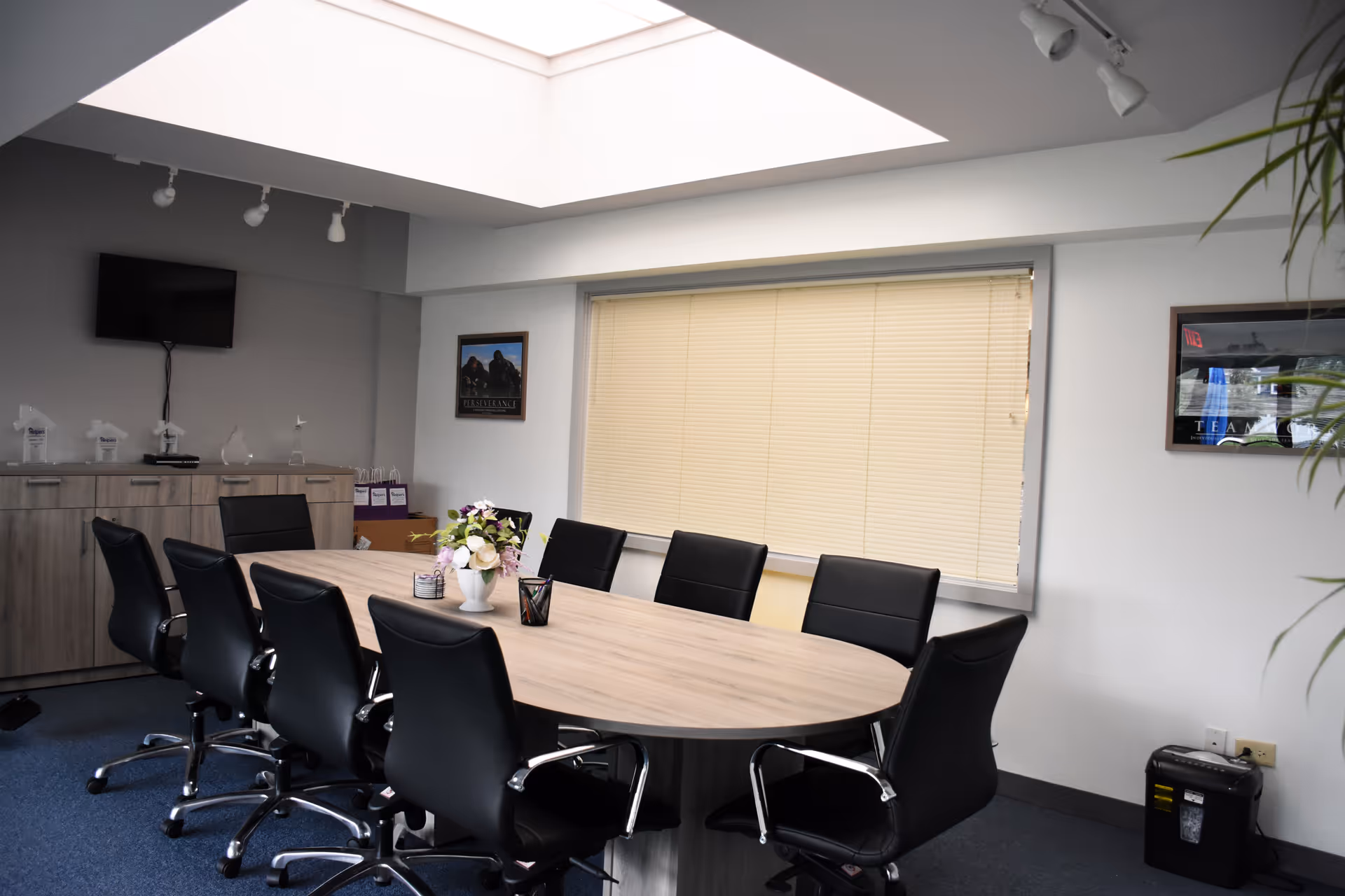 A modern conference room with a large oval table, black chairs, and window blinds.