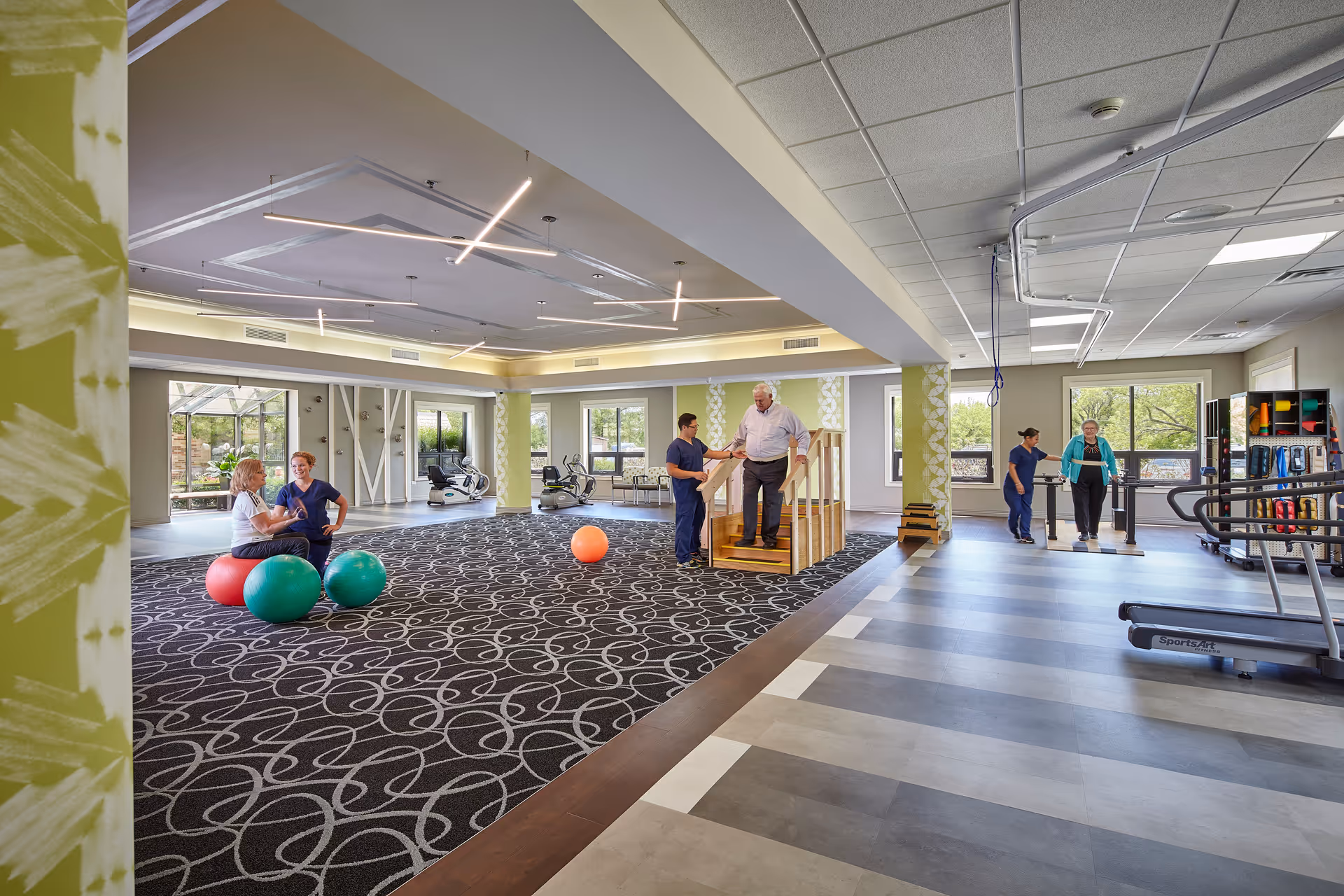 Spacious indoor rehab gym with exercise equipment, people exercising, and staff guiding residents.
