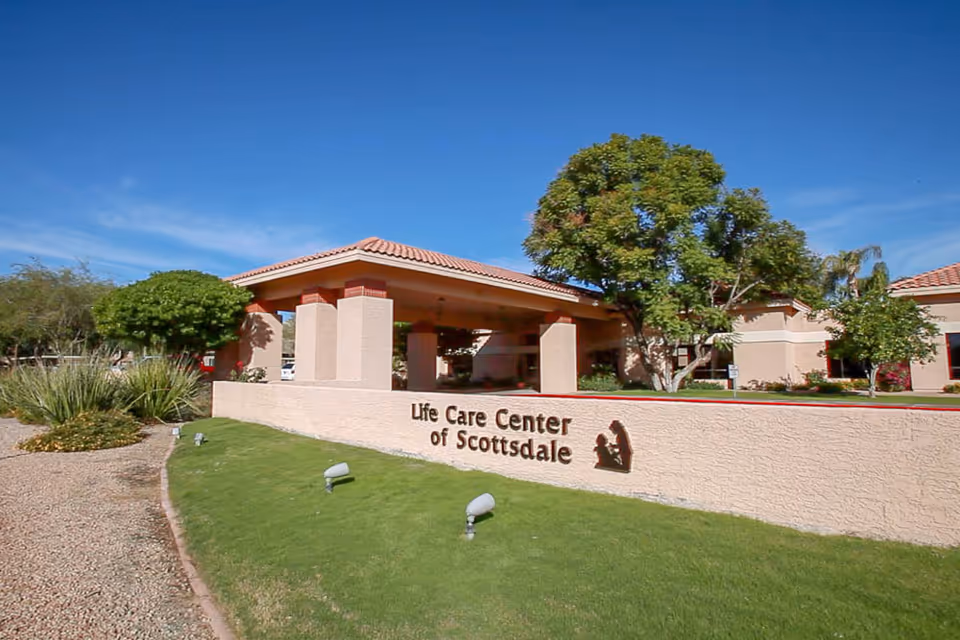 Front entrance of the Life Care Center of Scottsdale showing a low sign wall, covered porte-cochere, landscaping and a clear blue sky.