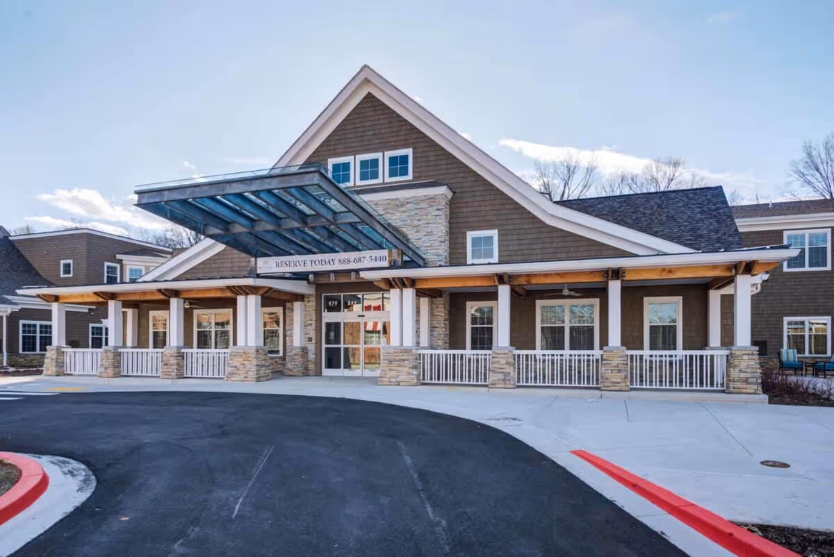 Front entrance of a multi-story senior living building with a glass-covered porte-cochere, stone columns, and a circular driveway.