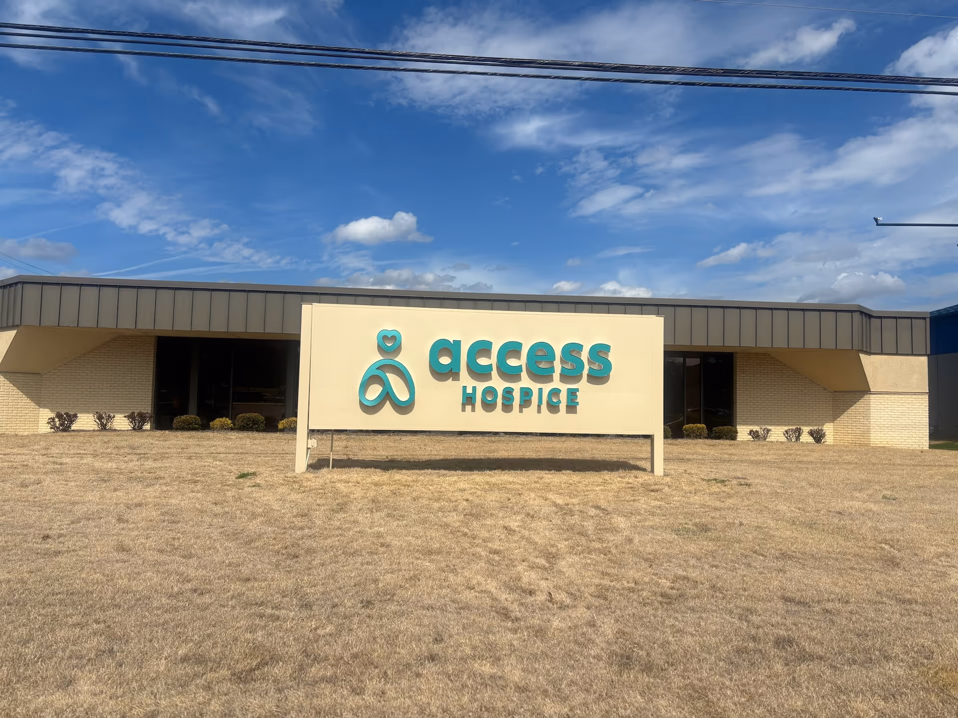 A single-story hospice building with a large teal Access Hospice sign on a dry front lawn.