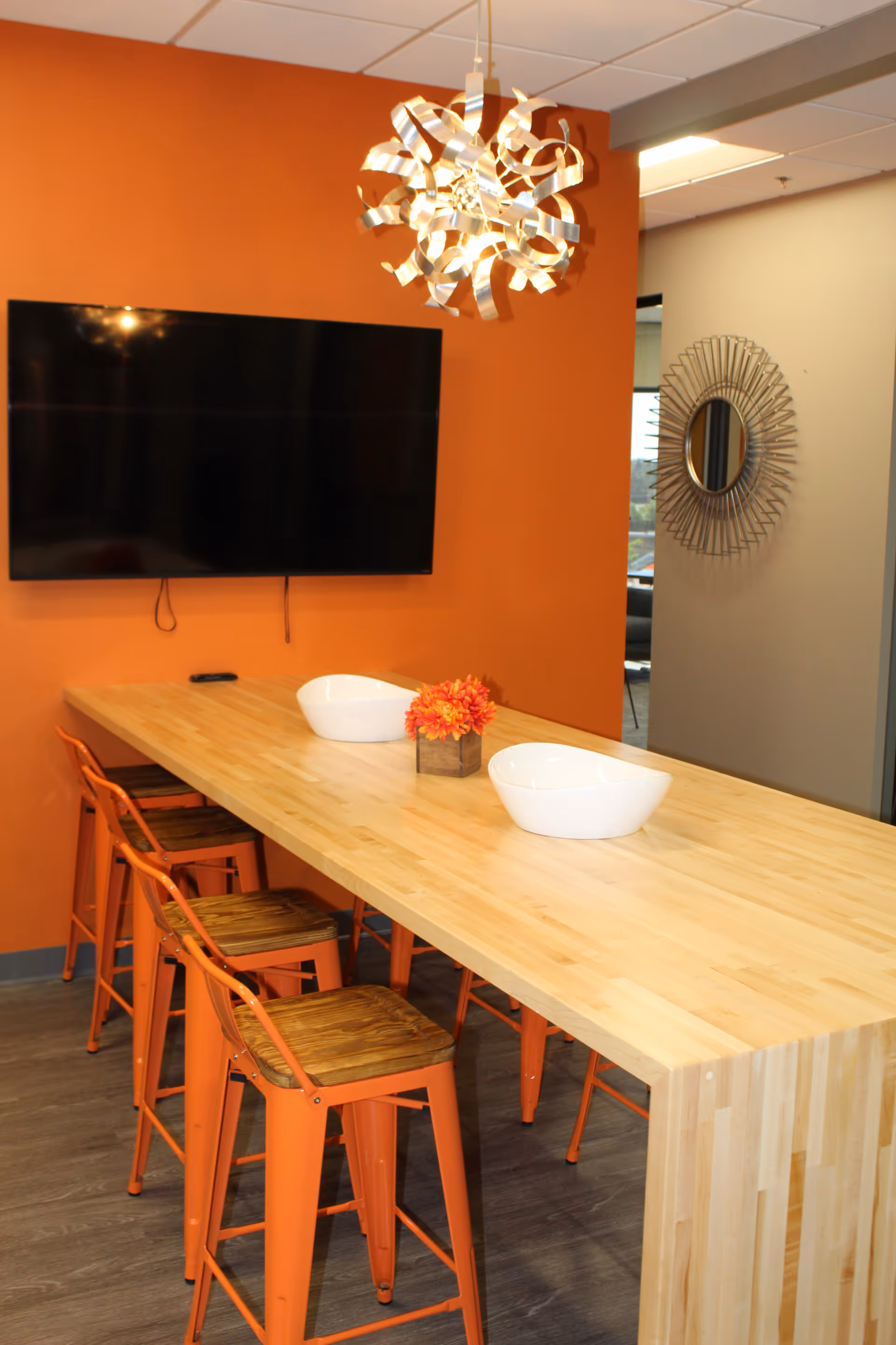 A bright dining area with a long wooden table, orange chairs, TV, and decorative lighting.
