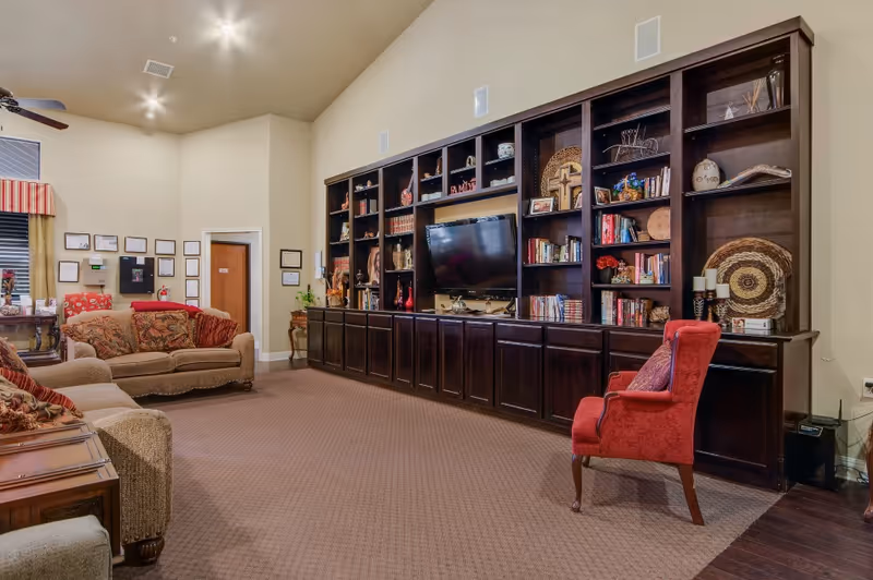 A spacious living room in a senior facility featuring a large dark wood bookshelf and seating.
