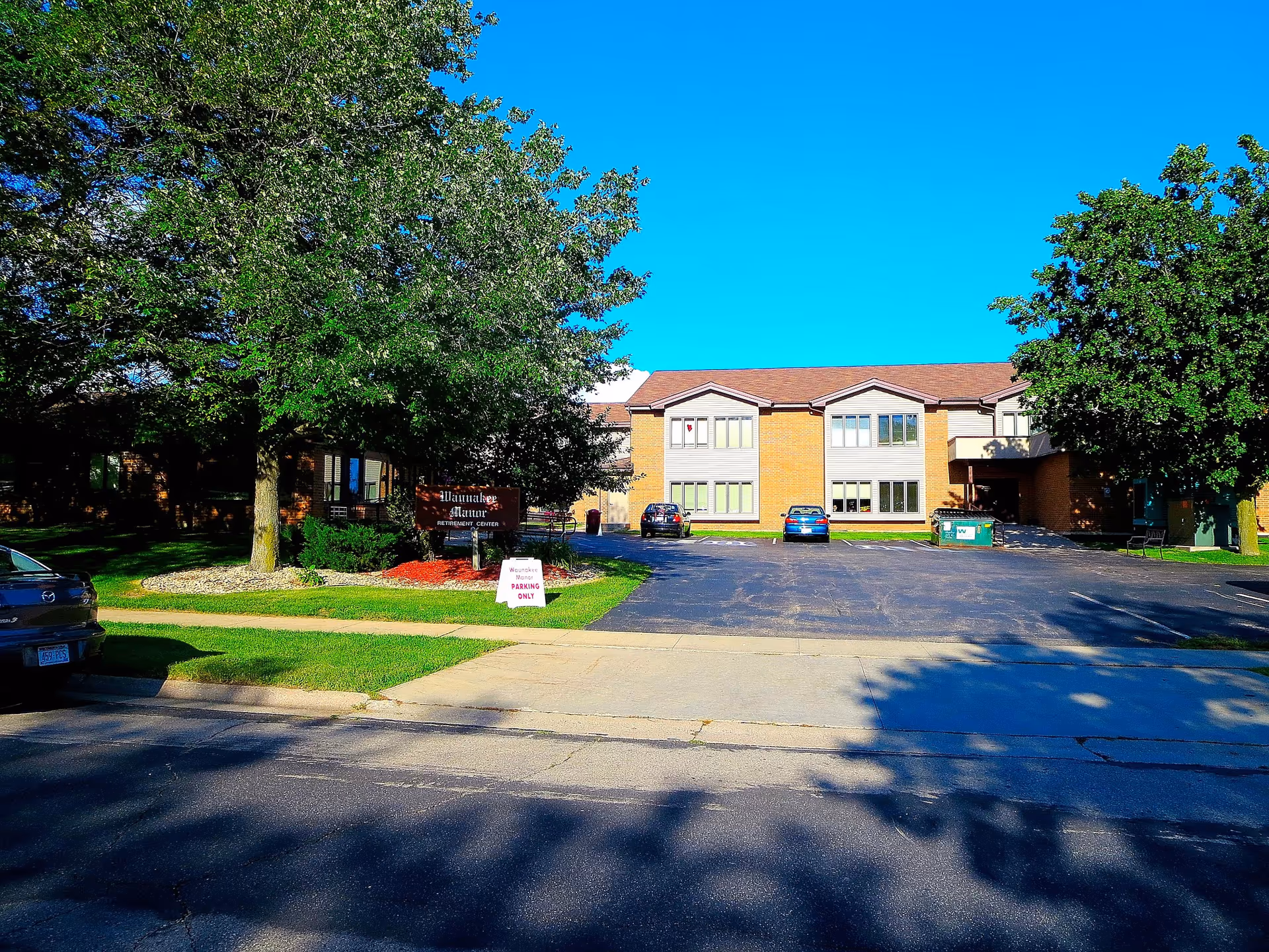 Exterior view of Waunakee Manor building with trees, parking lot, and blue sky