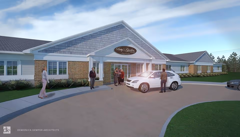 Exterior view of a single-story senior living facility with a covered entrance. Several people are standing near the entrance and a white car is parked in front. The building has a combination of brick and siding with a gabled roof. The sign above the entrance reads 'Betty's Garden'.
