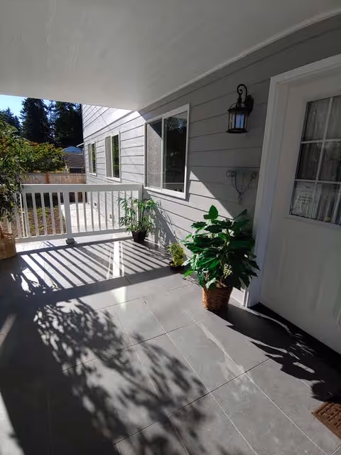 Shaded covered balcony with tiled floor, railing, exterior door, and potted plants.