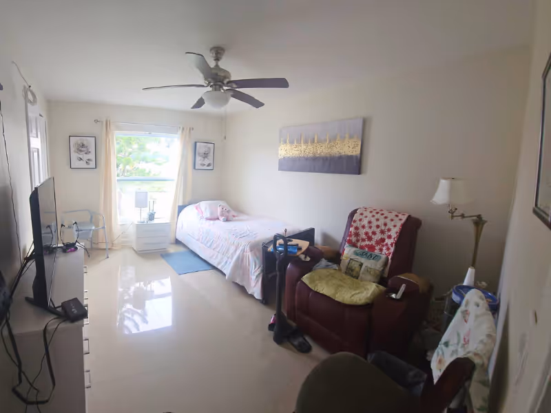 A small bedroom in a senior facility with a bed, recliner, TV, and ceiling fan