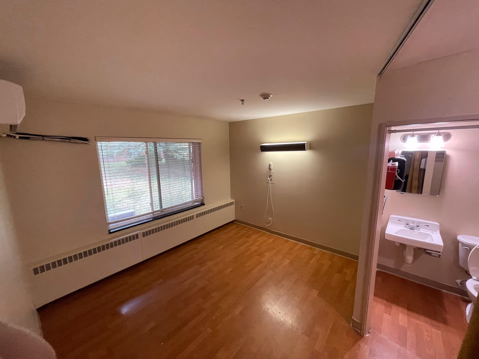 Empty senior living room with window, wood floor, wall light, and adjacent bathroom.