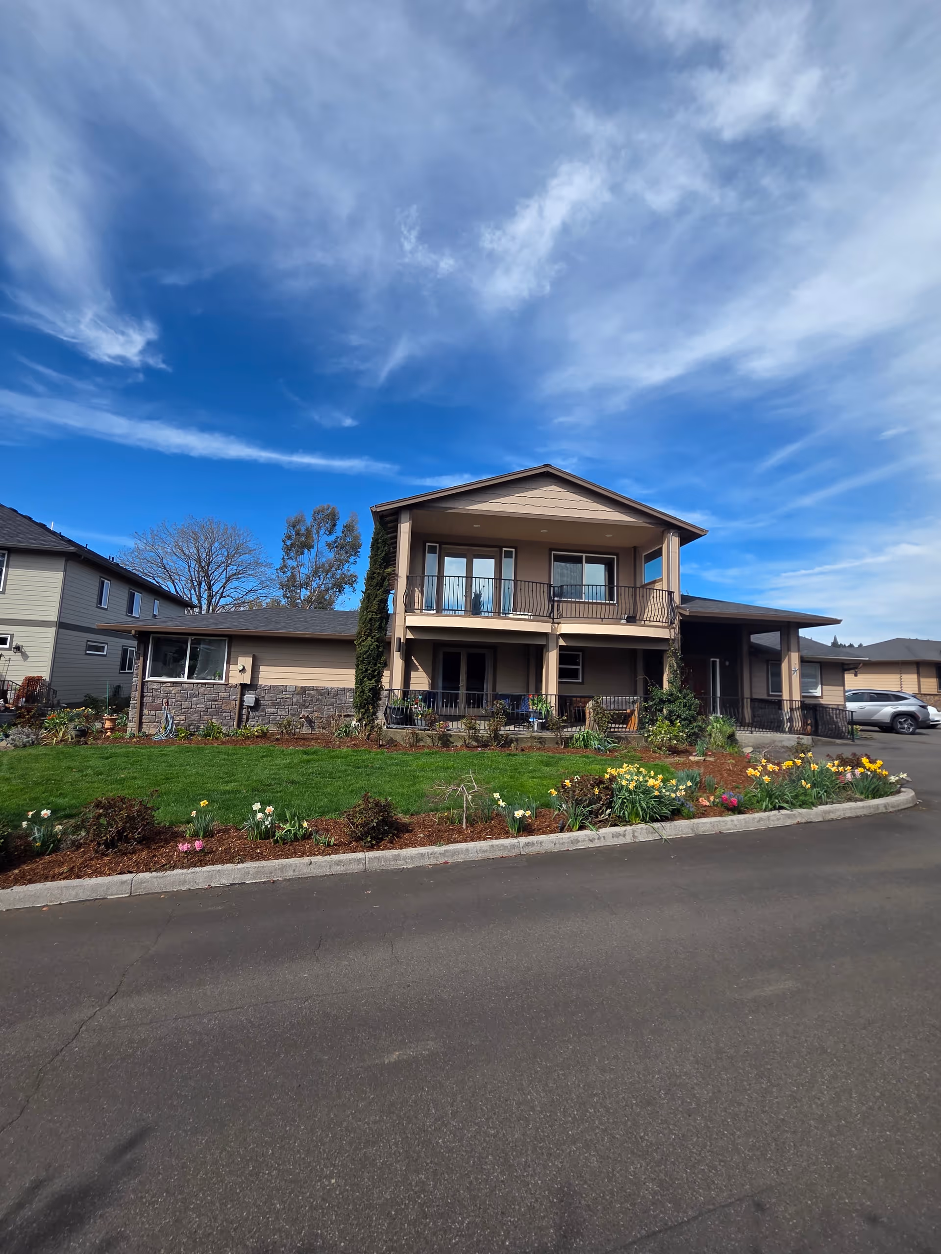 Two-story beige senior home with balcony, landscaped front yard, and blooming flowers.