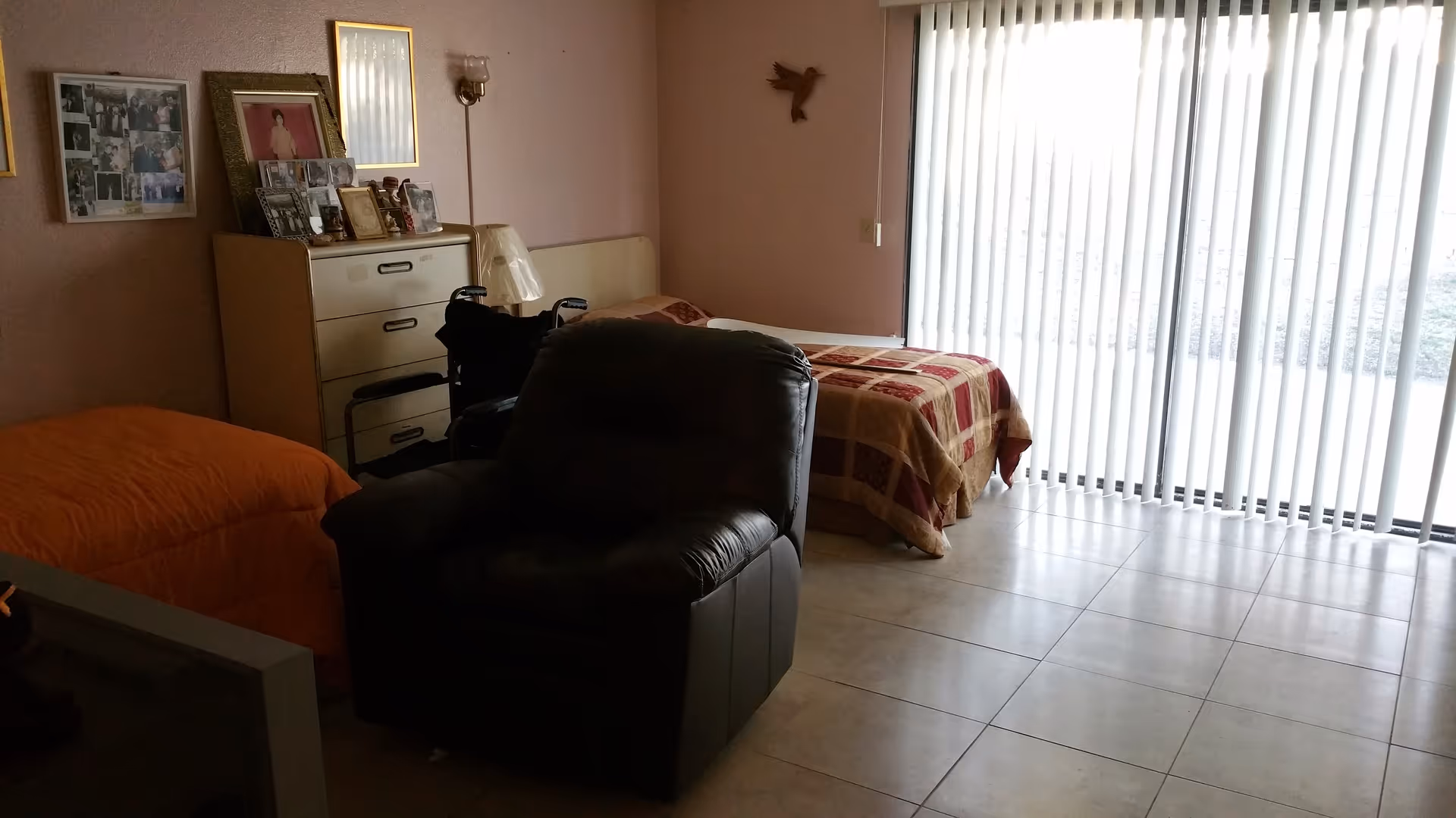 Indoor senior living bedroom with two beds, a recliner, dresser, photos, and sliding glass doors.