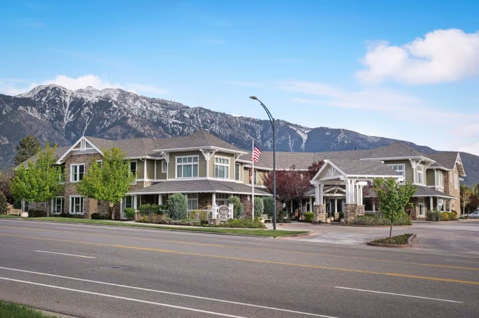 Exterior view of a two-story senior living facility building with a covered entrance, surrounded by trees and landscaping, with mountains in the background under a blue sky with some clouds.
