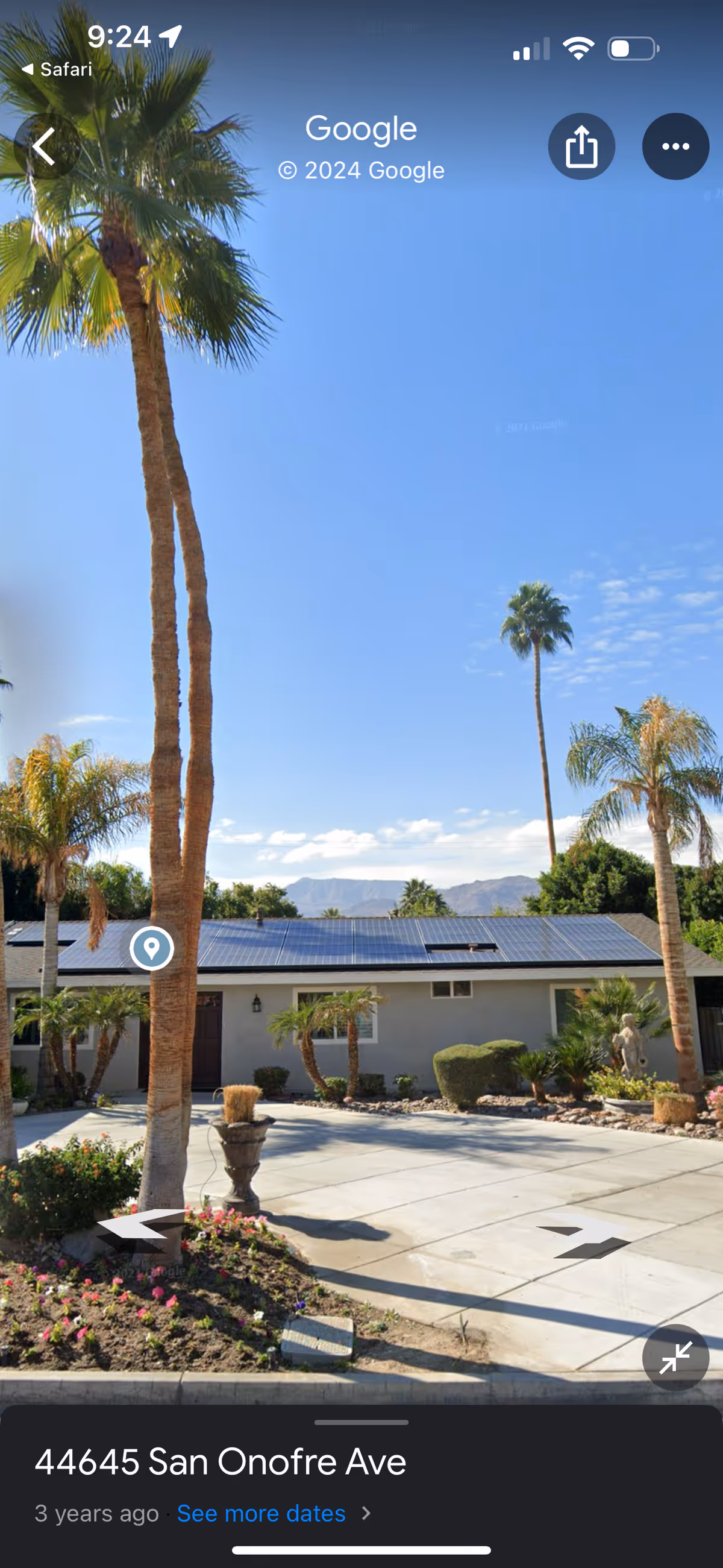 Exterior view of a single-story building with solar panels on the roof, surrounded by palm trees and landscaped plants under a clear blue sky with mountains in the background.