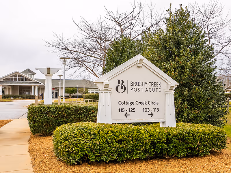 Outdoor view of the entrance sign for Brushy Creek Post Acute facility, surrounded by trimmed bushes and trees, with a building visible in the background under a cloudy sky.