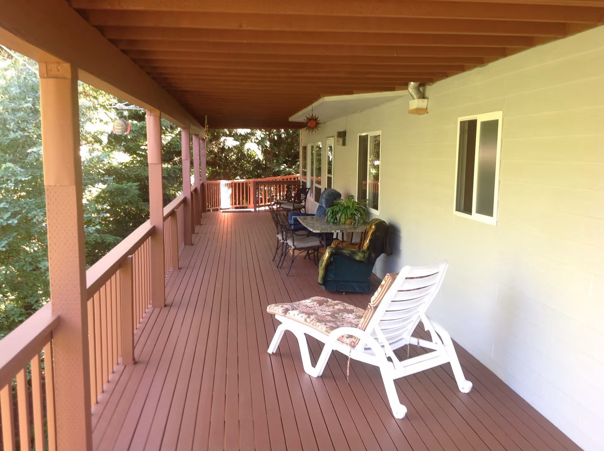 A covered outdoor wooden deck with several chairs, including a white lounge chair with a floral cushion, a table with chairs, and a recliner. The deck is surrounded by trees and has a railing along the edge.