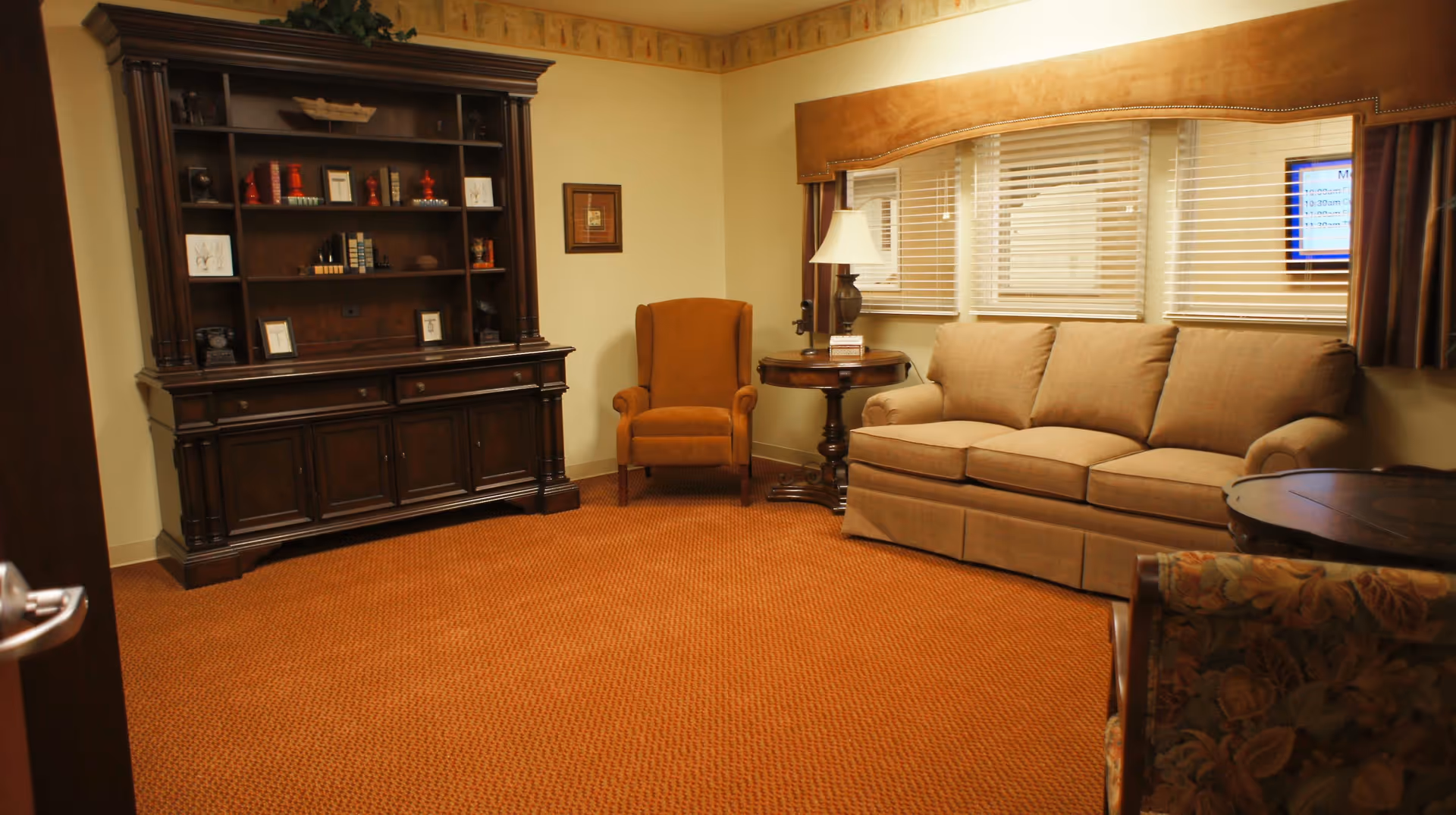 Cozy sitting room with a sofa, upholstered armchair, wooden cabinet and side tables beneath a window with blinds.