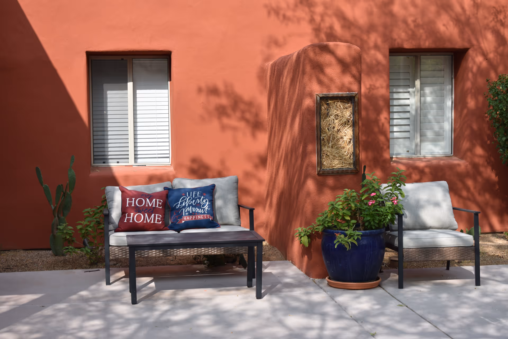Outdoor seating area with two cushioned chairs and a small table between them, placed against a terracotta-colored wall with two windows. One chair has two decorative pillows, one red with the words 'HOME HOME' and one blue with the words 'LIFE Liberty Pursuit HAPPINESS'. There is a large blue pot with green plants and pink flowers next to the chairs.