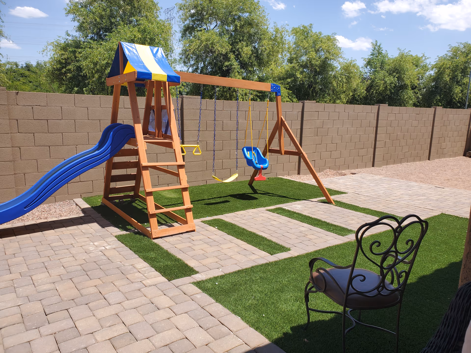 Outdoor play area with a wooden swing set that includes a blue slide, two regular swings, and a blue toddler swing. The ground is covered with artificial grass and stone pavers. There is a decorative metal chair with a cushion in the foreground. A tall brick wall and green trees are visible in the background under a clear blue sky.