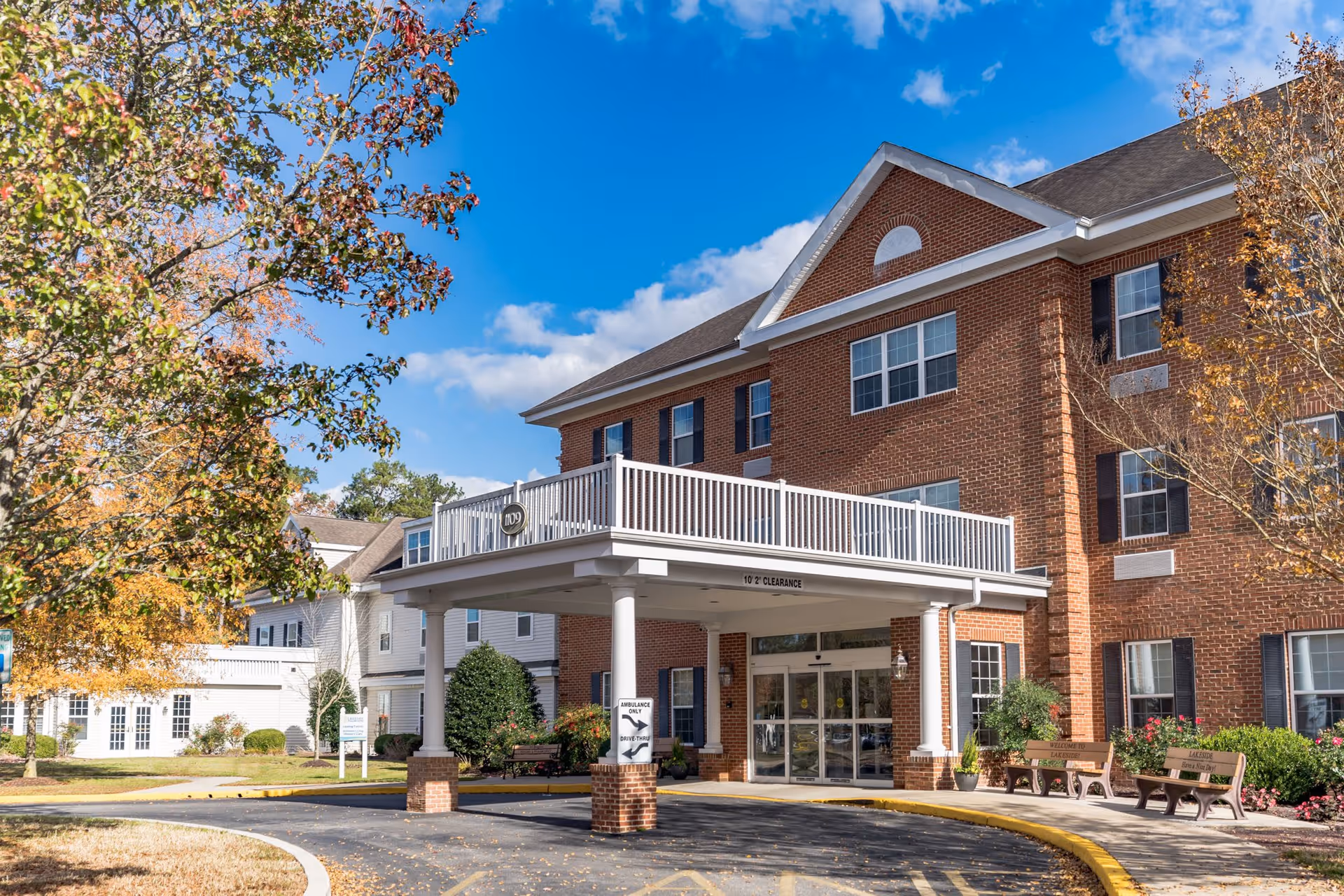 Exterior view of a senior living facility named Lakeside At Mallard Landing, featuring a brick building with multiple windows, a covered entrance with white pillars, benches, and landscaped greenery under a blue sky with some clouds.