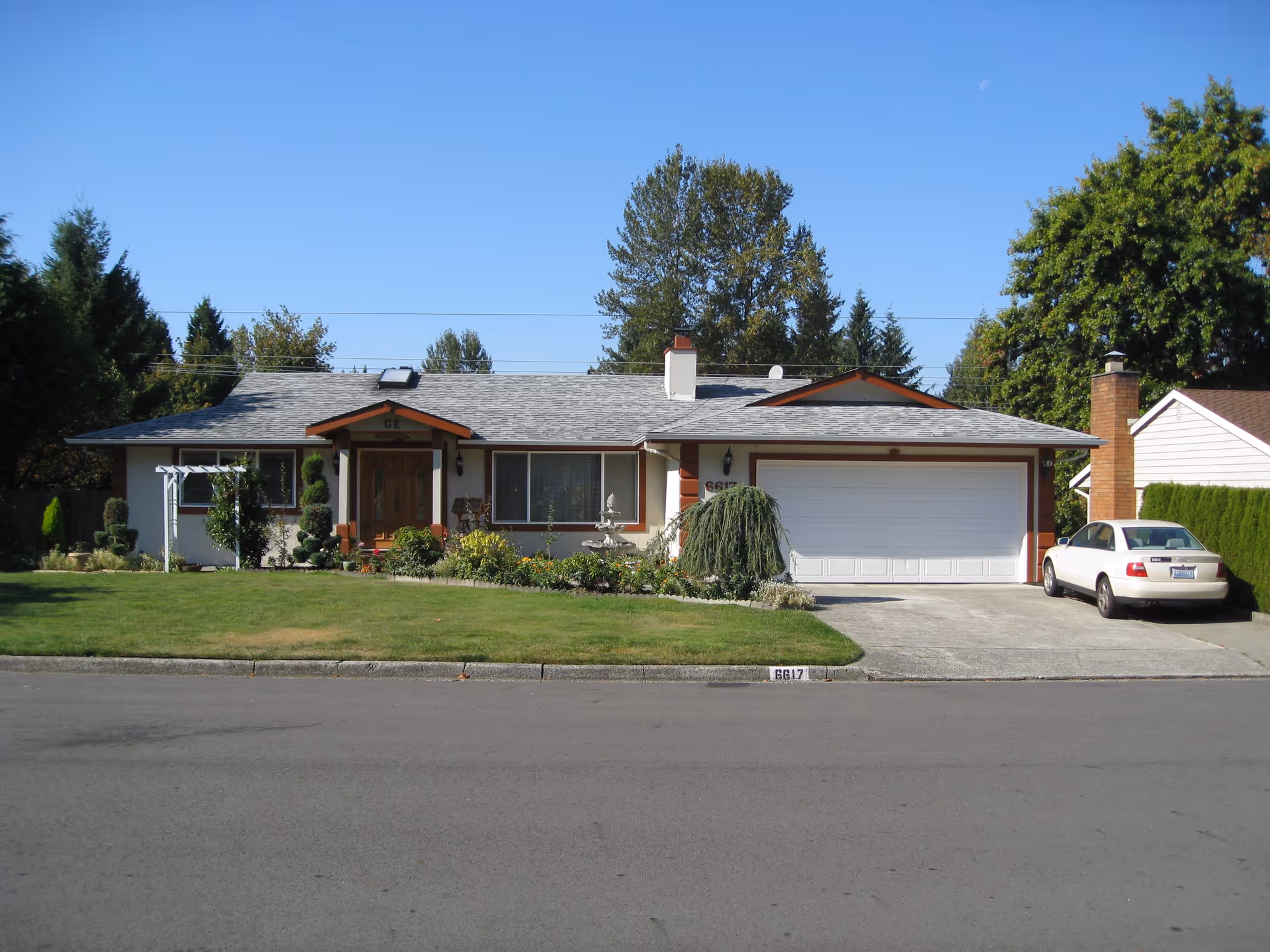 Single-story suburban house with a front lawn, attached garage, and a car parked in the driveway under a clear blue sky.