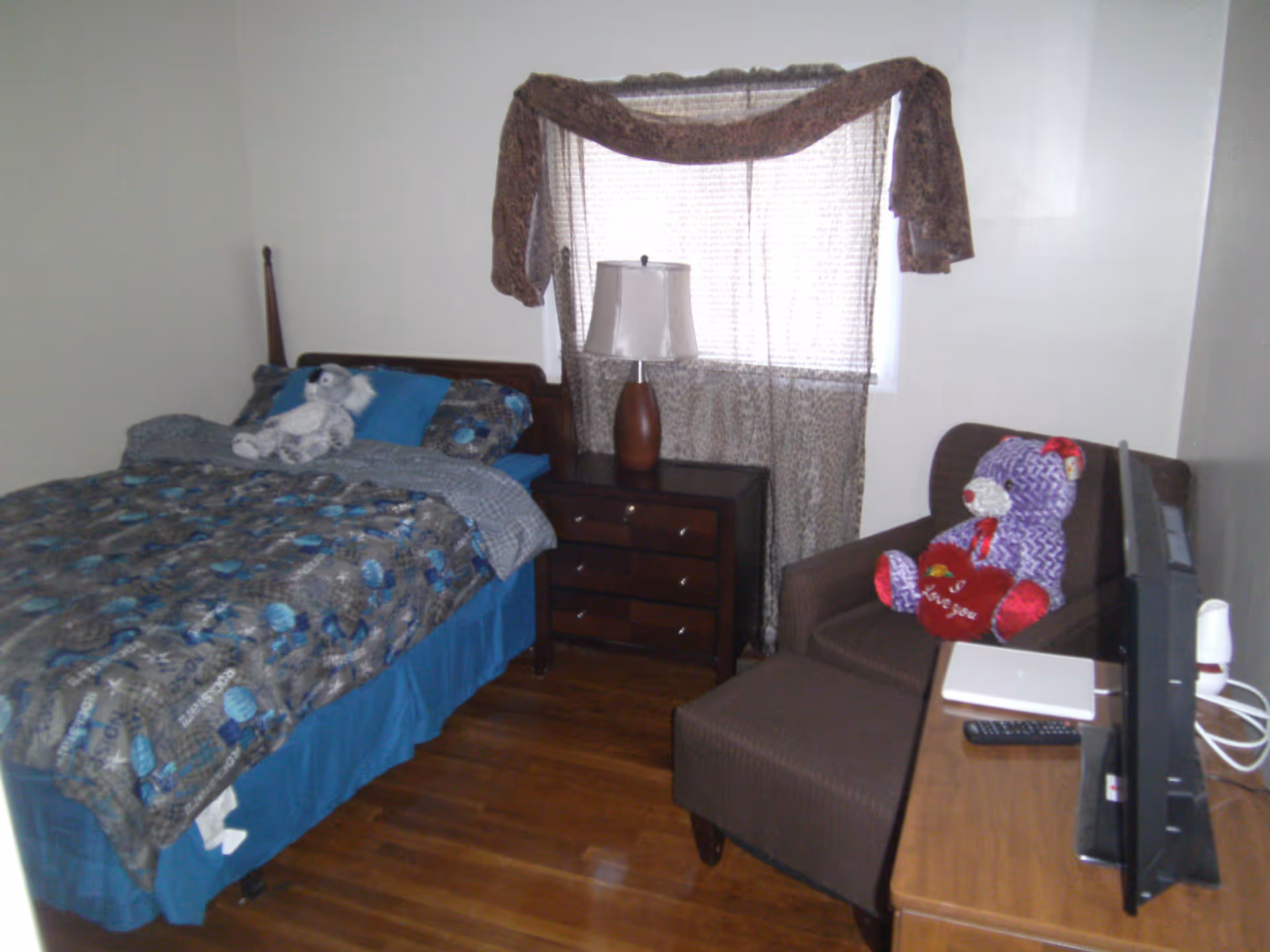 Small bedroom with a made bed, nightstand and lamp, an armchair with a teddy bear, and a TV on a dresser near a curtained window.