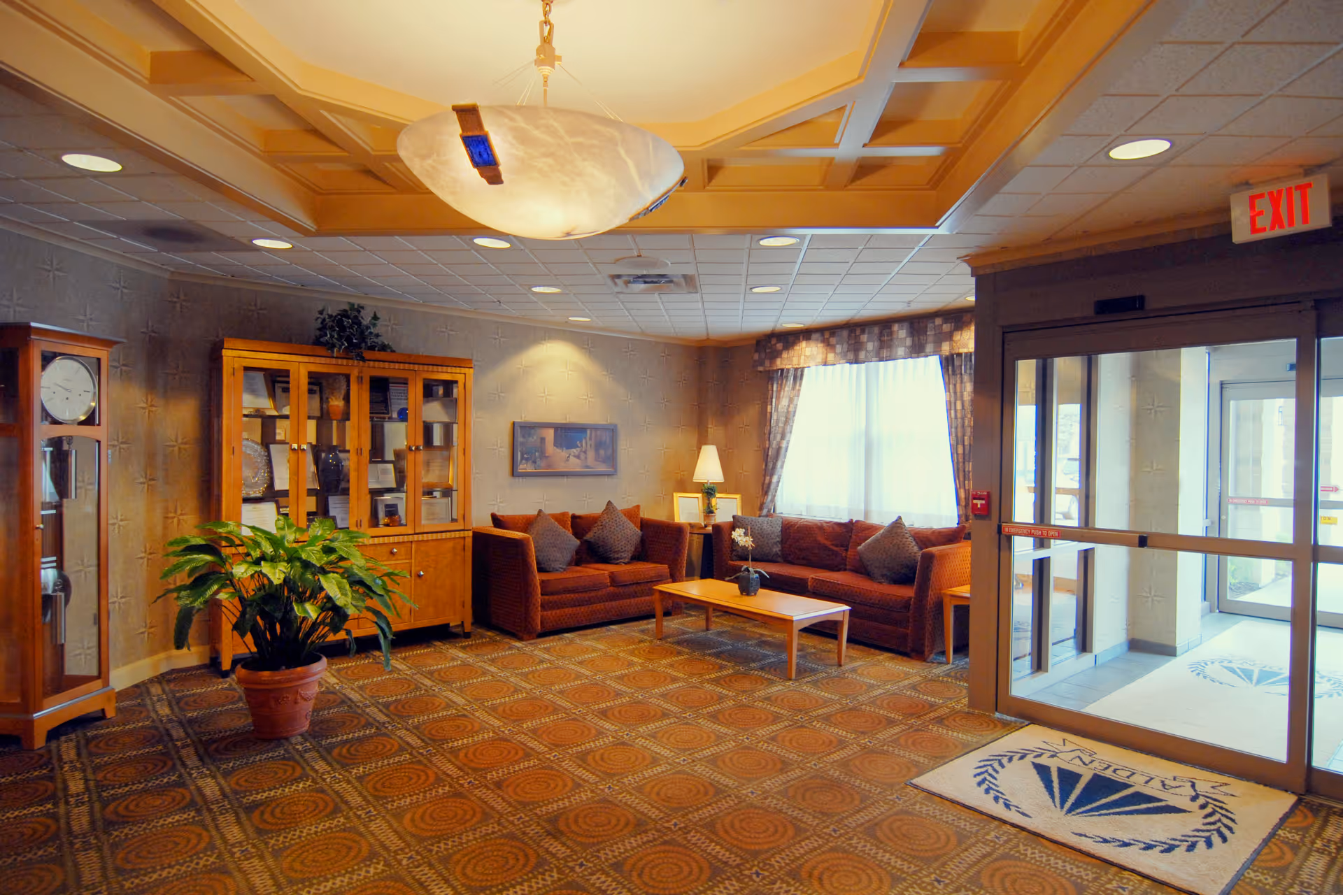 A cozy senior living facility lobby with two brown sofas, a wooden coffee table with a small plant, a wooden display cabinet, a grandfather clock, a potted plant, and a large window with curtains. The entrance door has glass panels and a mat with the Alden logo. The ceiling has recessed lighting and a decorative hanging light fixture.