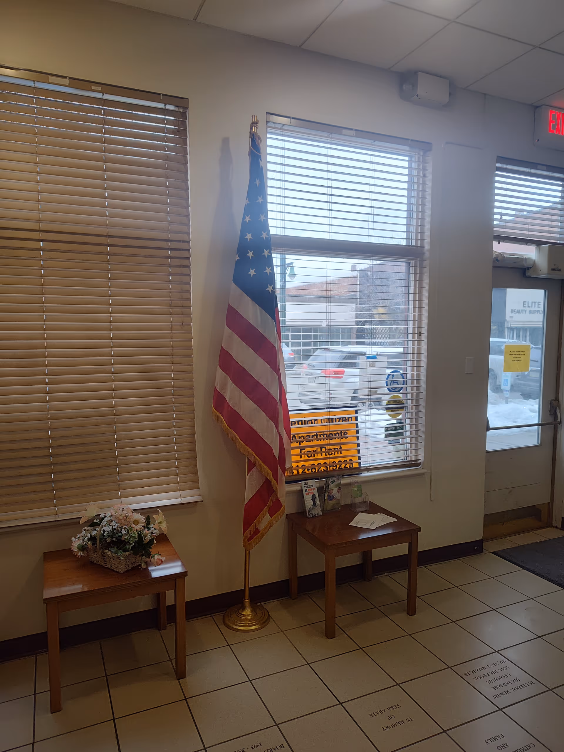 Interior lobby with American flag, two small tables, flower arrangement, and windows with blinds.