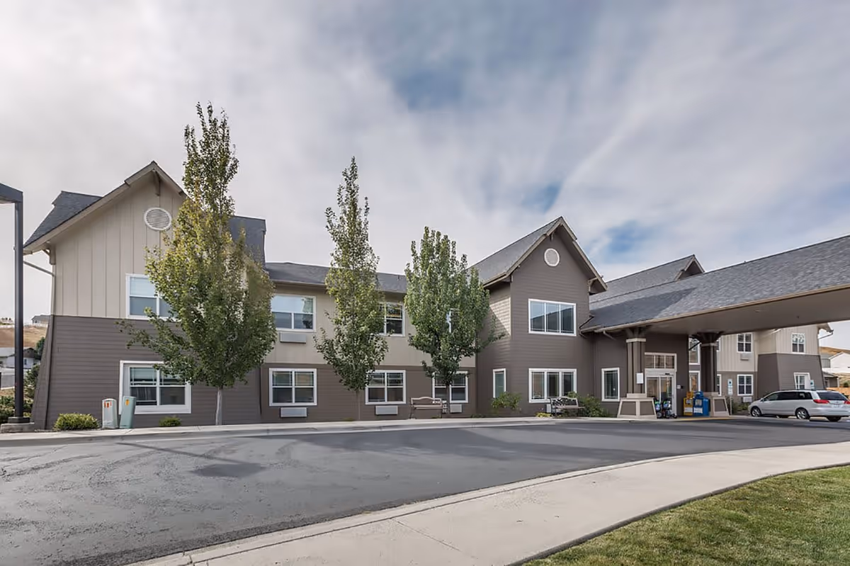 Exterior view of a two-story senior living facility building with beige and gray siding, several windows, and a covered entrance driveway. There are three tall trees in front of the building and a parked silver vehicle near the entrance. The sky is partly cloudy.