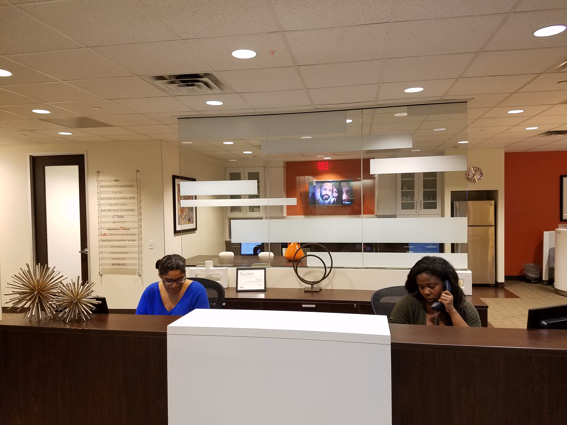 Two staff members sit at a dark wood reception desk behind glass partition in a lobby.